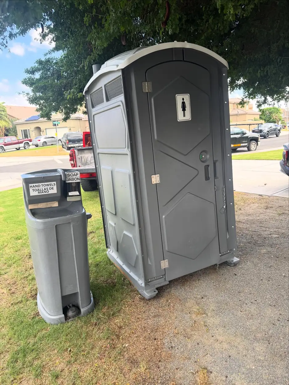 Gray porta potty with a foot-pump hand wash station beside it